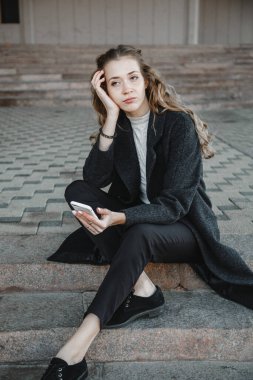 Siyah ceketli, kıvırcık saçlı bir kız elinde telefonla sokakta oturuyor.