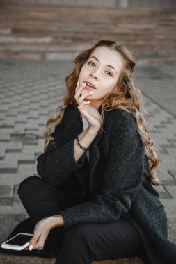 Kıvırcık saçlı kız siyah ceketli ve elinde telefonla sokakta oturuyor.