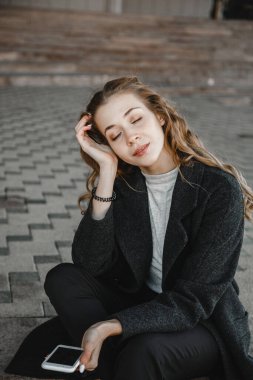 Kıvırcık saçlı kız siyah ceketli ve elinde telefonla sokakta oturuyor.