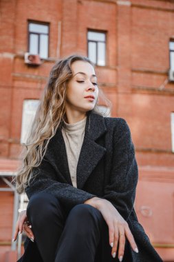 Siyah ceketli kıvırcık kız kırmızı bir binanın arkasında poz veriyor.