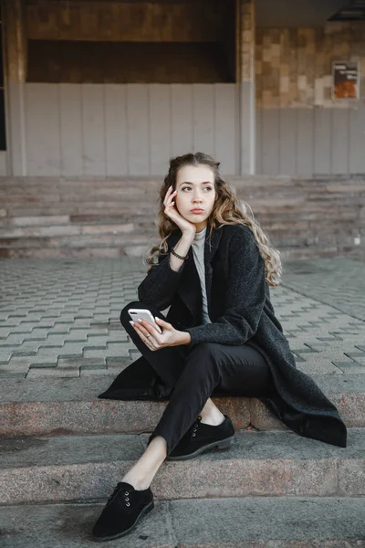 卷曲头发的女孩穿着黑色外套坐在街上 手里拿着电话 — 图库照片