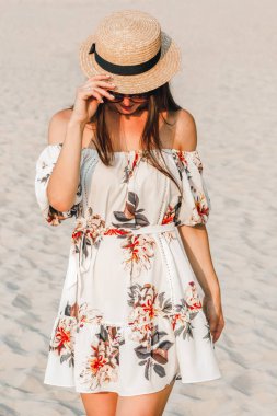 Kumsalda poz veren beyaz elbiseli, şapkalı ve gözlüklü bir esmer.