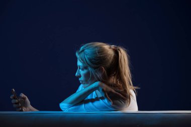 Güzel, uzun saçlı sarışın kadın oynuyor, göz gezdiriyor, cep telefonuna bir kanepenin üzerinde oturuyor, mavi ve doğal renk ışıklarıyla aydınlanıyor, koyu mavi arka planda..