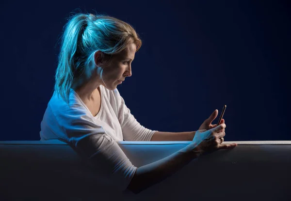 美丽的金发女人在沙发上玩耍 看她的手机 被蓝色和自然的彩灯照亮 背景是深蓝色的 免版税图库图片