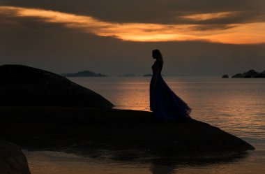 Günbatımı, plaj, uzun elbise standında bir kayanın üzerinde ince kadın.