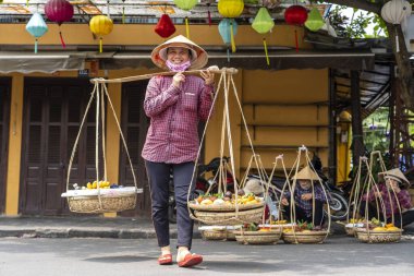 Hoi An, Vietnam - 16 Mart 2020: Vietnam 'daki Hoi An, Vietnam' daki eski sarı duvarın arka planında elinde meyve sepeti olan hasır şapkalı Vietnamlı kadın