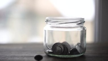 Pencerenin yanındaki tahta masada cam kavanoza düşen Ukrayna sikkeleri, kapat. Yavaş çekimde, gümüş para yığını arka plandaki boş bir cam kavanoza düşer. Ukrayna