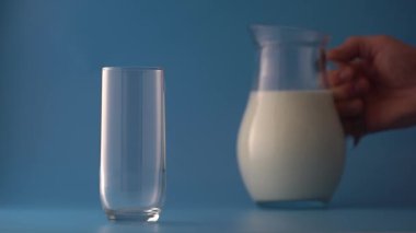 Süt bir sürahiden mavi arka plandaki boş bir bardağa dökülür, yakın plan. Süt ürünü konsepti