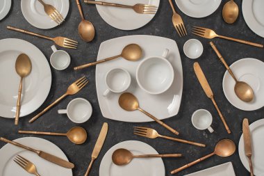 Birçok beyaz seramik tabak, fincanlar ve pirinç çatallar, bıçaklar ve siyah arka planda kaşıklar. Üst görünüm, kapat
