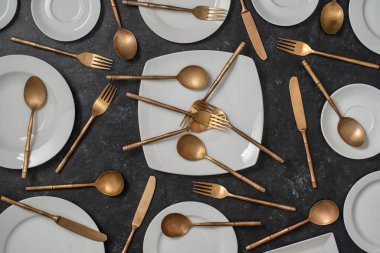 Siyah arka planda bir sürü beyaz seramik tabak, pirinç çatal, bıçak ve kaşık var. Üst görünüm, kapat