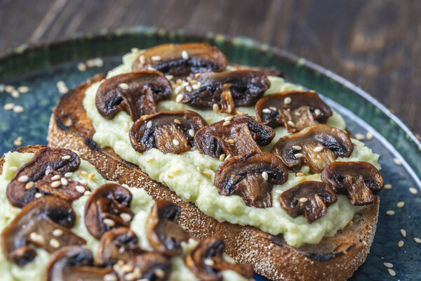 Tasty sandwich with green mashed avocado, fried mushrooms and sesame seeds in plate on a table, close up. Vegetarian food