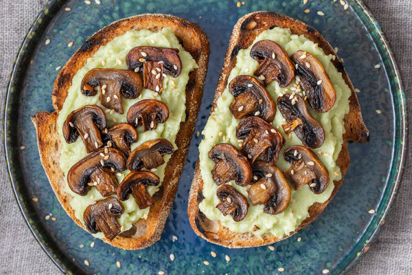 Tasty sandwich with green mashed avocado, fried mushrooms and sesame seeds in plate on a table, close up. Vegetarian food