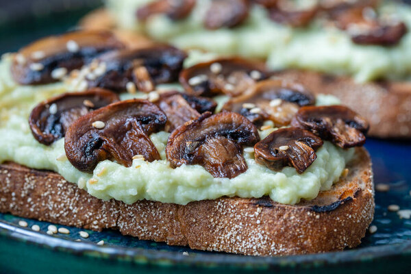 Tasty sandwich with green mashed avocado, fried mushrooms and sesame seeds in plate on a table, close up. Vegetarian food