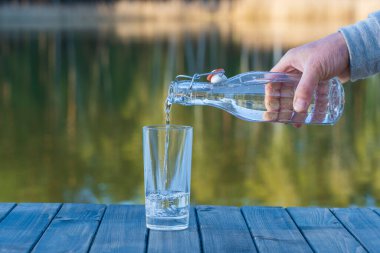 İlkbaharda göl ve ormanın yanındaki bir şişeden bardağa su dökmek. Kapan. Doğa ve seyahat kavramı