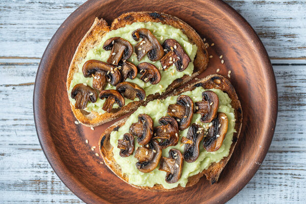 Tasty sandwich with green mashed avocado, fried mushrooms and sesame seeds in plate on a table, close up. Vegetarian food