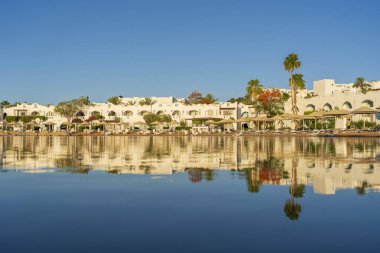 Binalar, güneş panelleri, şemsiyeler ve palmiye ağaçları, Mısır, Afrika 'daki Sharm El Sheikh tatil beldesinde sabahleyin sahildeki sakin Kızıl deniz suyuna yansıyor. Karantina sırasında boş plajlar