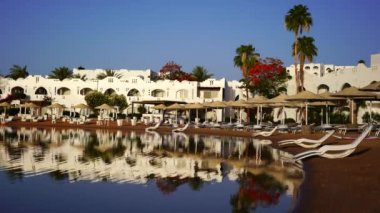 Gündoğumunda, Mısır 'ın Sharm El Sheikh tatil beldesindeki sakin deniz suyuna binalar, güneşlikler ve şemsiyeler yansıtılır.