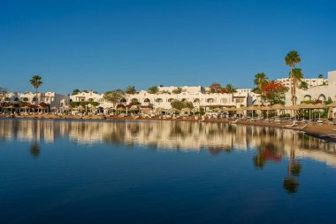 Binalar, güneş panelleri, şemsiyeler ve palmiye ağaçları, Mısır, Afrika 'daki Sharm El Sheikh tatil beldesinde sabahleyin sahildeki sakin Kızıl deniz suyuna yansıyor. Karantina sırasında boş plajlar