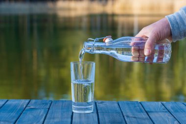 İlkbaharda göl ve ormanın yanındaki bir şişeden bardağa su dökmek. Kapan. Doğa ve seyahat kavramı