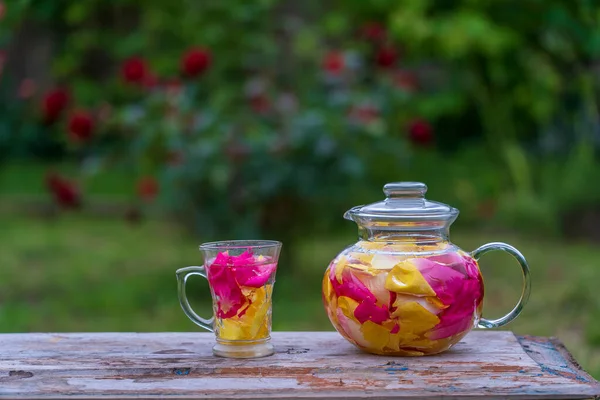 木製のテーブルの夏の庭で ピンク 赤の花びらのバラから美しく香りの良いお茶がグラスティーポットとマグカップにバラ 自然を背景にバラの花びらからハーブティーを閉じます — ストック写真