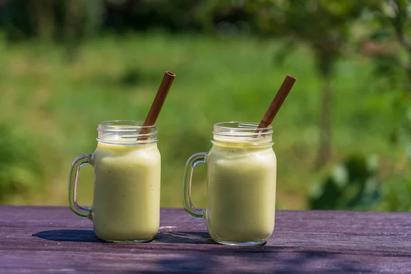 Fresh green smoothie from avocado, banana and honey in glass mug on wooden table in nature background, close up. Concept of healthy eating. Green smoothie in sunny summer day in garden