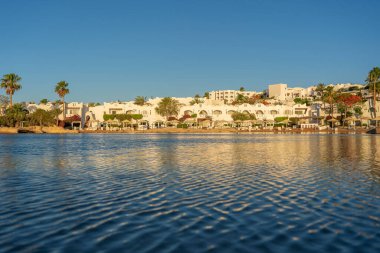 Binalar, güneş panelleri, şemsiyeler ve palmiye ağaçları, Mısır, Afrika 'daki Sharm El Sheikh tatil beldesinde sabahleyin sahildeki sakin Kızıl deniz suyuna yansıyor. Karantina sırasında boş plajlar