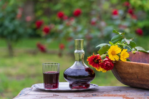 木製のテーブルの上に夏の庭の背景に花の花びらで飾られた赤ワインとガラスのデカンタとガラス クローズアップ — ストック写真