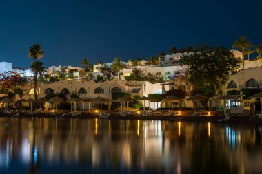 Binalar, güneş panelleri ve şemsiyeler Mısır 'ın Sharm El Sheikh tatil beldesindeki kumsaldaki sakin deniz suyuna yansıtılır.