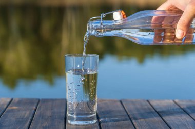 İlkbaharda göl ve ormanın yanındaki bir şişeden bardağa su dökmek. Kapan. Doğa ve seyahat kavramı