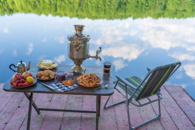 Ukrayna 'nın yeşil ormanlarında sakin su gölü yakınındaki masada beyaz duman ve yiyecek bulunan klasik metal çay semaverleri. Doğa gıda konsepti