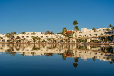 Binalar, güneş panelleri, şemsiyeler ve palmiye ağaçları, Mısır, Afrika 'daki Sharm El Sheikh tatil beldesinde sabahleyin sahildeki sakin Kızıl deniz suyuna yansıyor. Karantina sırasında boş plajlar