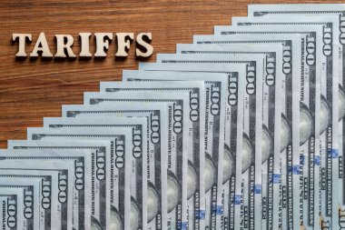 Üzerinde yazı tarifeleri ve masa üzerinde 100 dolarlık kağıt banknotlar olan ahşap harfler. Trump yönetimi sırasındaki ticaret savaşlarını ve gümrük vergilerini sembolize eden gümrük vergileri ABD dolarlarının üzerine bindi