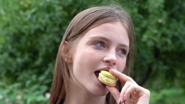Çekici, gülümseyen genç sarışın kız badem kurabiyelerine bakar ve onu yer, doğa arka planı. Gençlik duyguları, açık hava, yakın plan portre