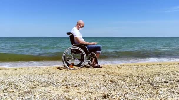 Une personne handicapée portant un masque lit un livre au bord de la mer assise dans un fauteuil roulant — Video