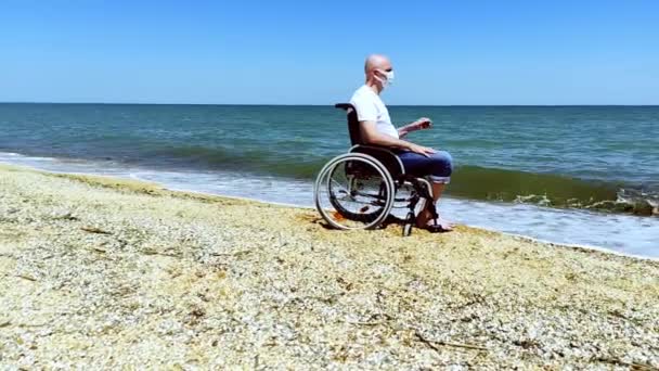 Gehandicapte in een rolstoel doet het masker af en ademt frisse lucht in aan zee — Stockvideo