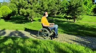 Maskeli engelli kişi parkta tekerlekli sandalyede otururken müzik dinliyor.