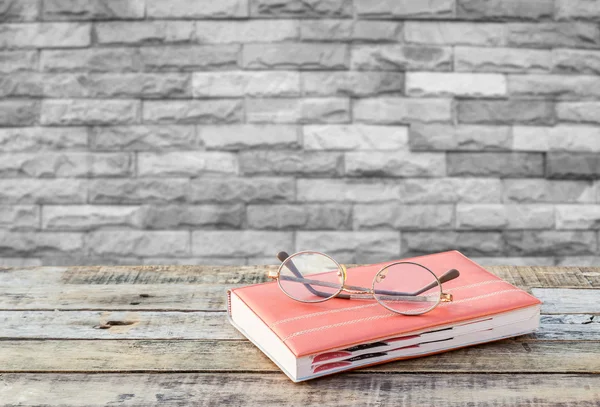 Notebook and eyeglasses on wooden table with blurred brick wall — Stock Photo, Image