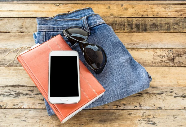Notebook,sunglasses,smart phone and blue jeans on wooden table — Stock Photo, Image