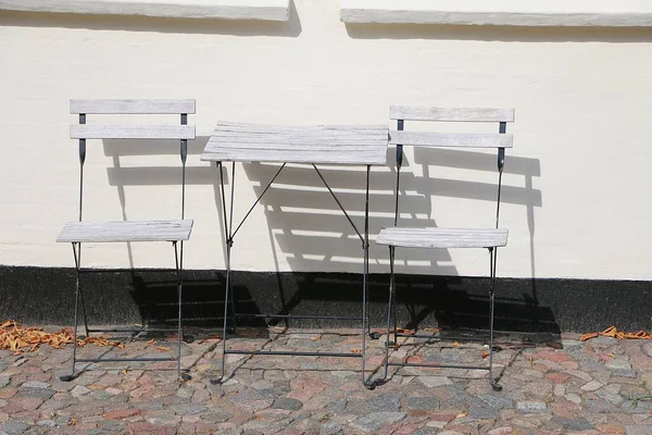 İki beyaz katlanır sandalye ve küçük beyaz katlanır bir masa güneşin altında evin duvarında duruyor.