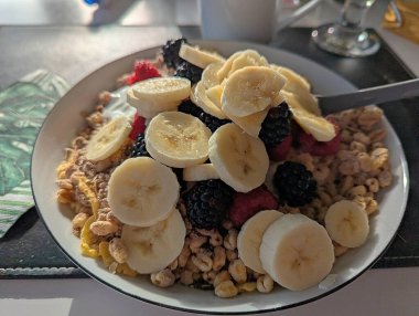 Taze meyve, muz dilimleri, ahududu, böğürtlen ve böğürtlen ile kaplı bir tabak dolusu müsli masada yemeye hazır..
