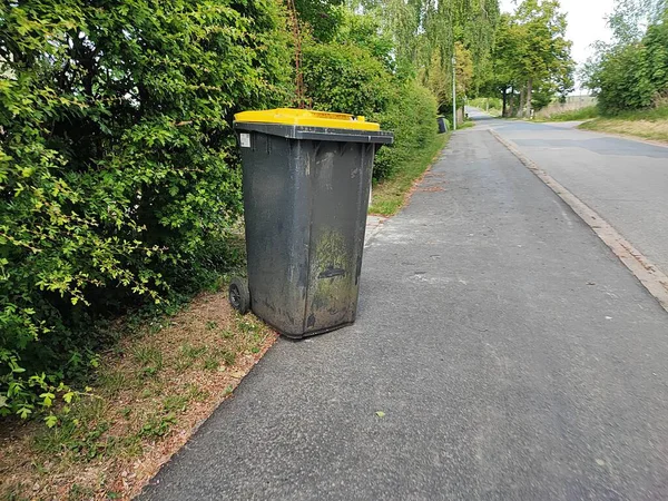 Sarı kapaklı siyah bir çöp tenekesi kaldırımdaki çitin yanında toplanmaya hazır bir şekilde duruyor.