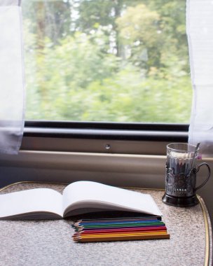 Tren kompartımanında bir çizim defteri, boya kalemleri ve cam bir masa.