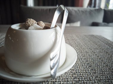 Beyaz, koyu ve siyah şeker ve gri sırtında metal cımbız olan cam bir kase.