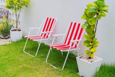 İki kırmızı ve beyaz plaj sandalyesi çimenlerde ve evin bahçesinde beyaz beton bir duvara karşı dizilir..