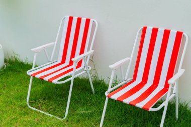 İki kırmızı ve beyaz plaj sandalyesi çimenlerde ve evin bahçesinde beyaz beton bir duvara karşı dizilir..