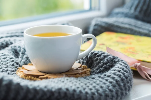 Weiße Tasse Kamillentee mit grauem Schal auf der Fensterbank. — Stockfoto