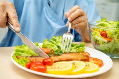 Hastanede sağlıklı yiyecekler için somon kazığı ve sebze salatası yiyen Asyalı yaşlı kadın hasta..