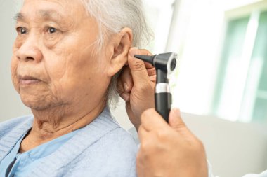 Odiyolog veya ENT doktoru otoskop kullanarak işitme sorunu olan Asyalı yaşlı kadın hastaların kulaklarını kontrol eder..