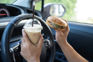 Arabada yemek için hamburger ve buzlu kahve taşıyan Asyalı kadın tehlikeli ve kaza riski taşıyor..