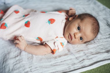 Yeni doğmuş bebek kameraya bakıyor. Evde battaniyeye uzanmış güzel kıyafetler giyiyor.